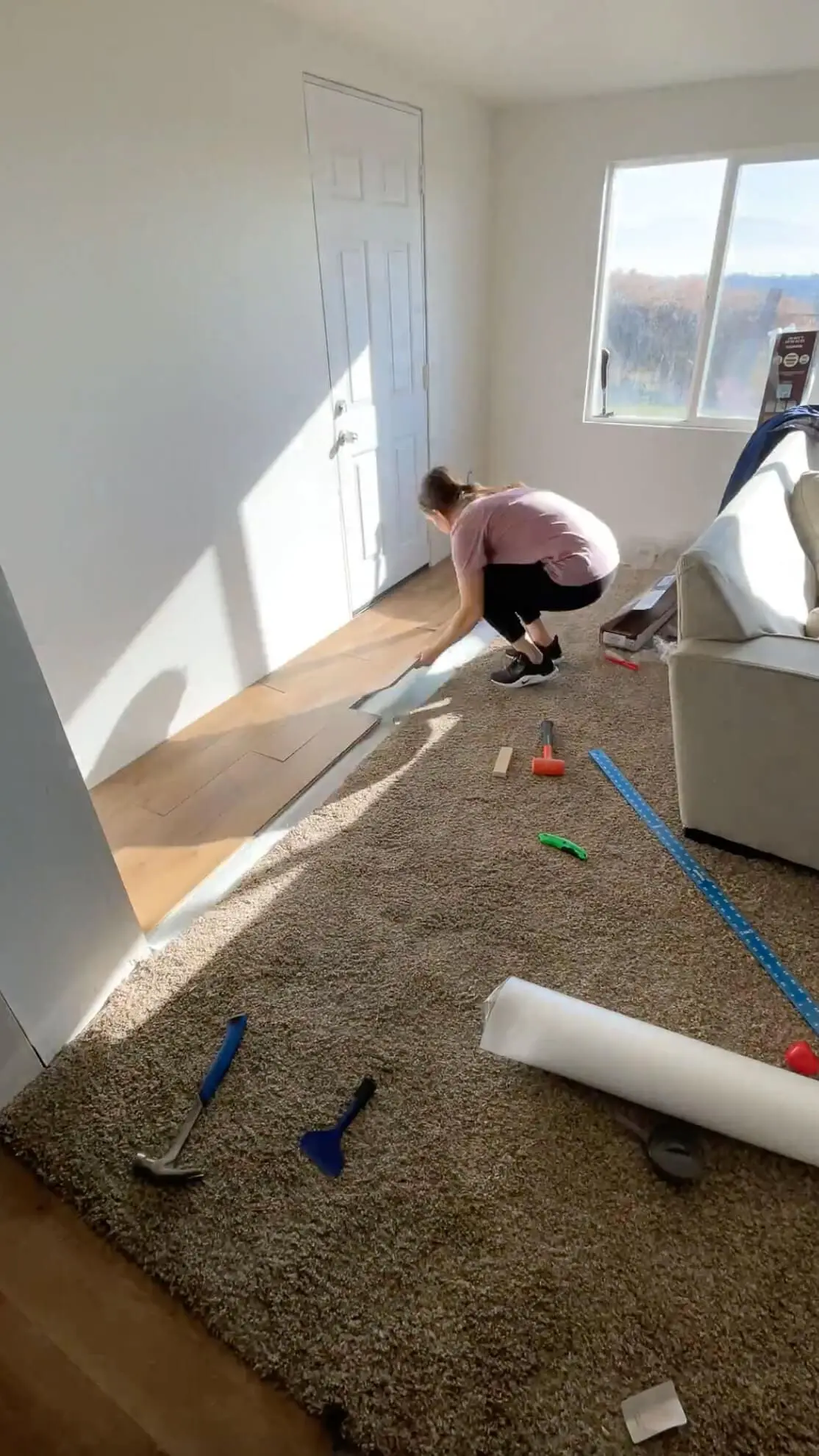 A woman installing wood-look flooring over a newly prepared surface near a white door. The room is filled with natural light, and various tools like a hammer, scraper, and measuring tape are visible on the carpeted area.